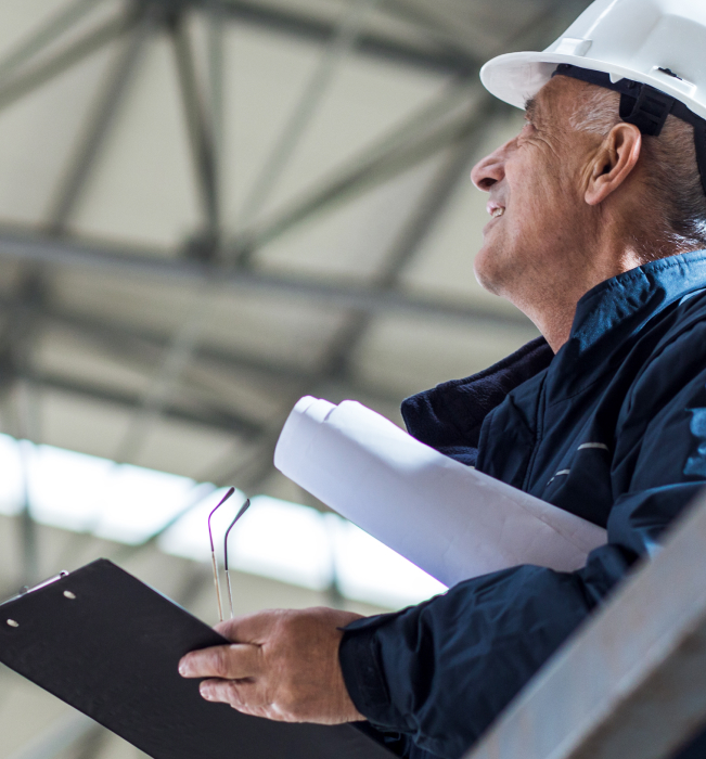 closeup image of a man in a hardhat holding glasses, plans and a clipboard looking into the roof of a warehouse